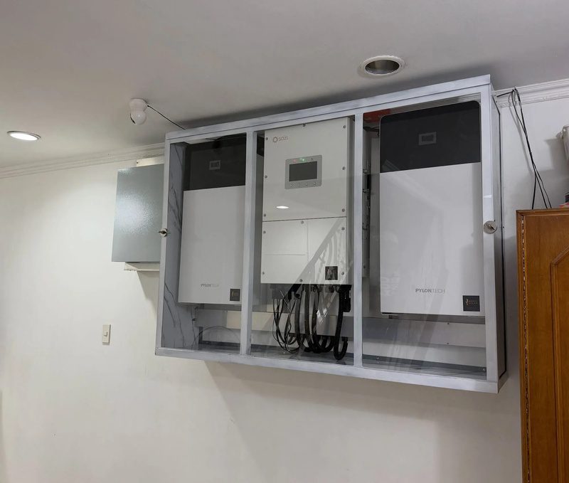 A hybrid inverter between two wall batteries inside a glass-fronted metal cabinet, mounted high on a living-room wall.