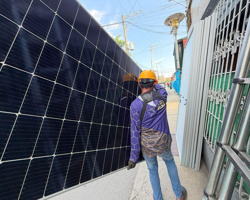 An installer in a hard hat, seen from behind, walking a full-size solar panel down a narrow street to the job.