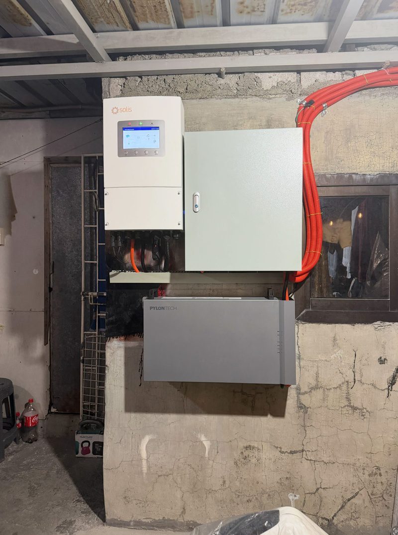 A hybrid inverter, a protection box and a wall battery on a garage wall, orange conduit running up to the roof.