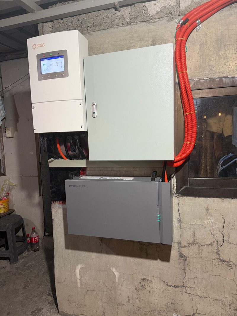 Close view of the hybrid inverter beside its protection box, the wall battery mounted below, conduit bundled above.