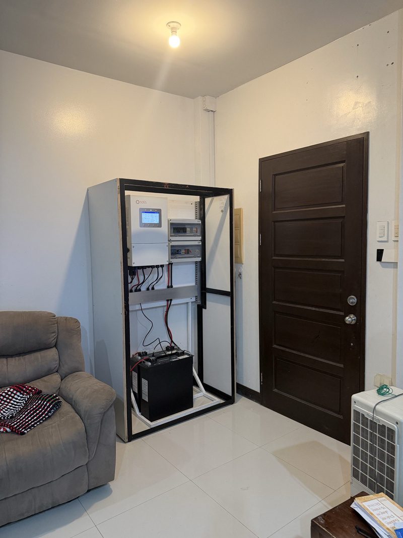 The open cabinet in the living room corner, inverter screen on, cables dressed to the battery.